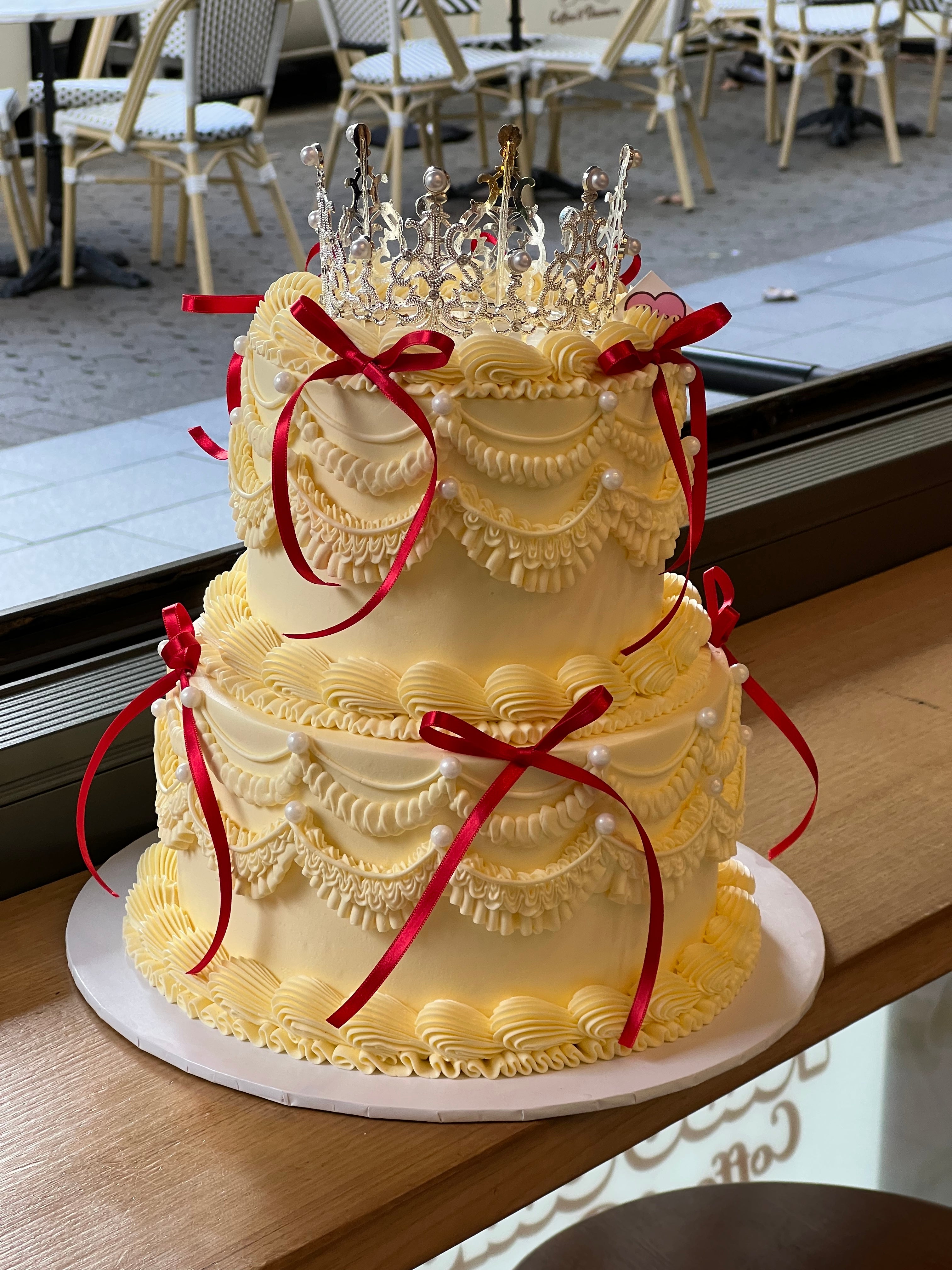 Two-tier vintage-style cream cake decorated with red ribbons, pearl piping, and a silver crown toppe