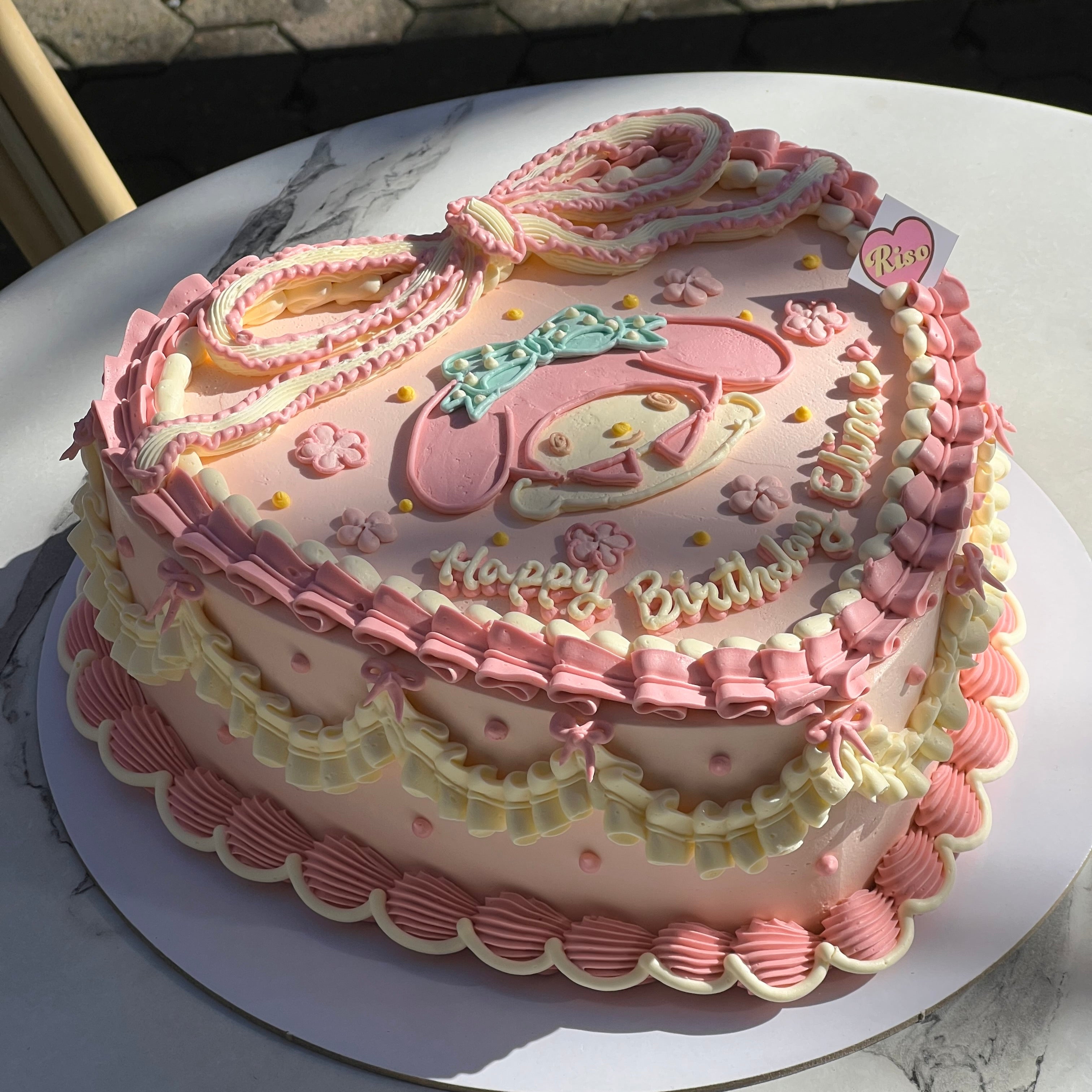 A pink vintage-style cake featuring a My Melody character and large piped ribbon on top, decorated with frilly borders and cream ruffles