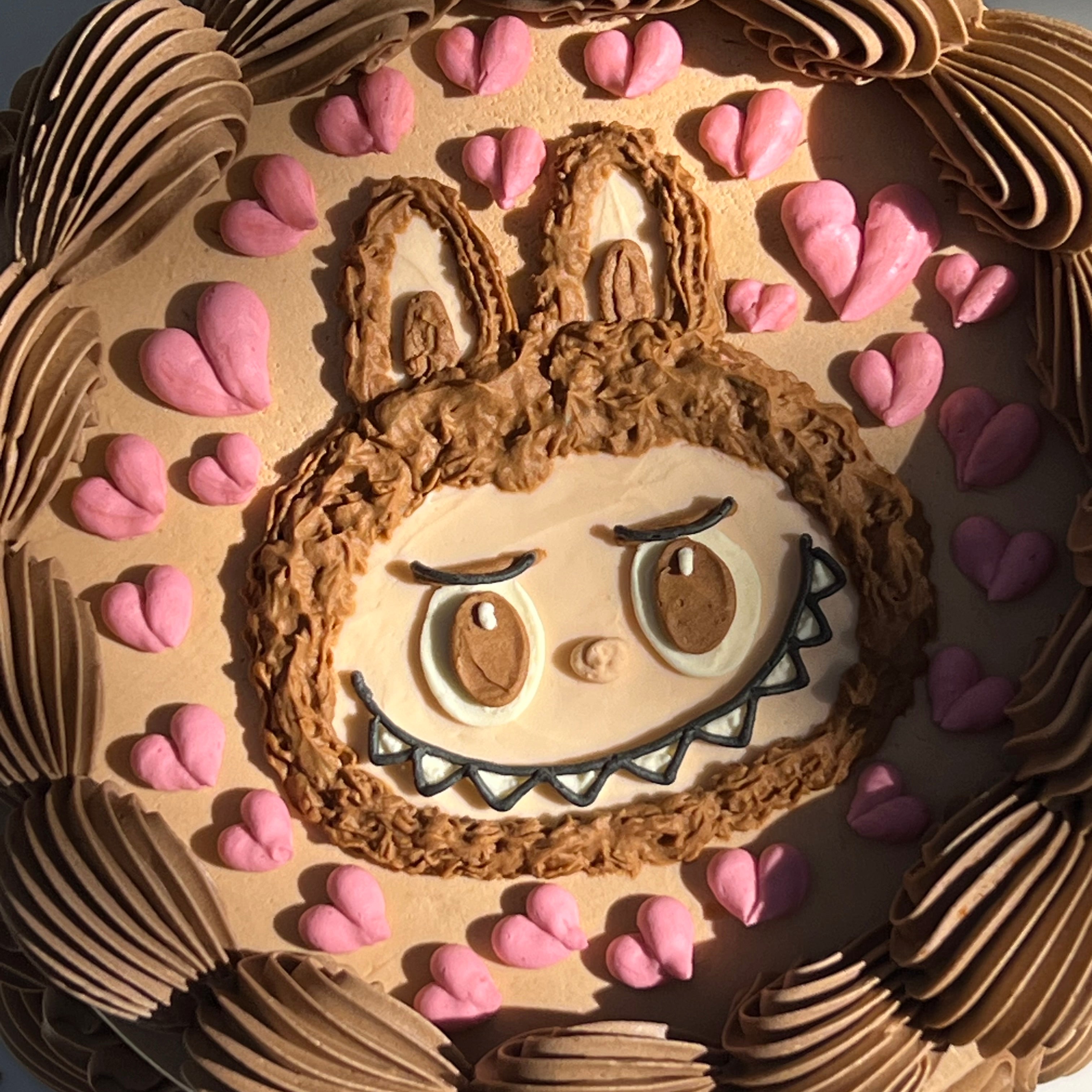Detailed view of a Labubu face cake with thick brown piping and scattered pink hearts, in a round shape