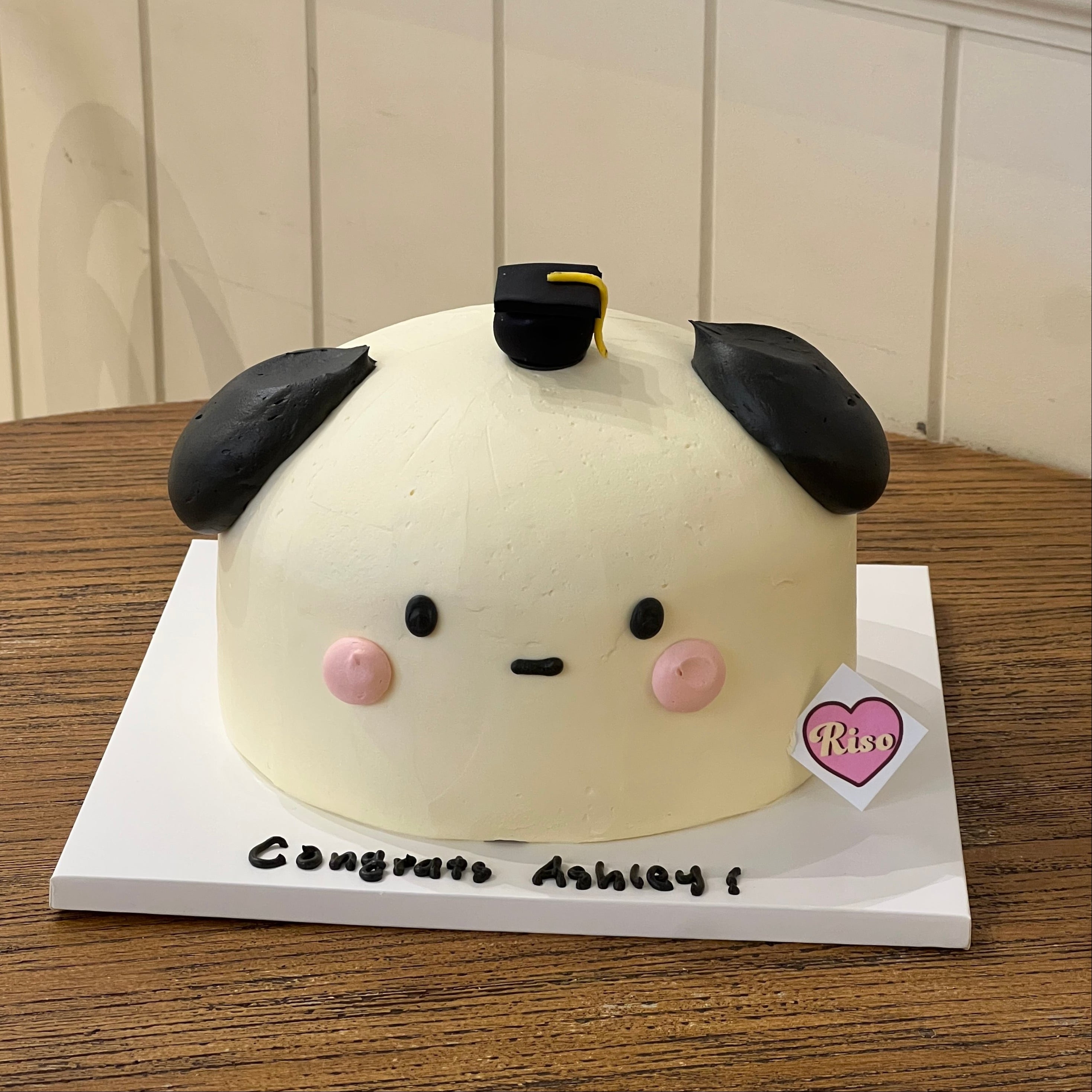 Graduation celebration cake with marshmallow-style design, decorated with a mini black cap and pink cheeks