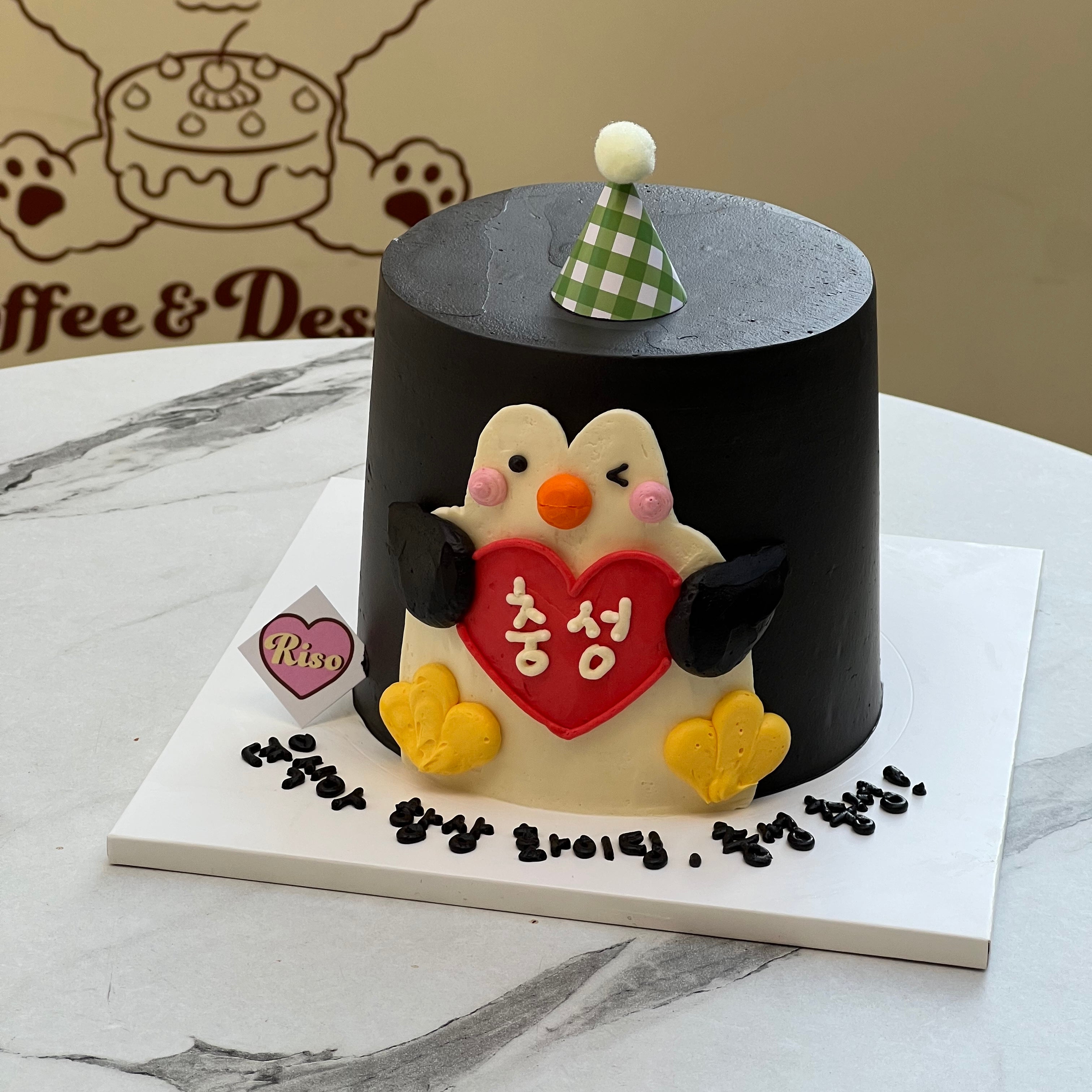 Top-down view of a minimal black cake with a 3D penguin topper and green party hat, showcasing clean lines and standout character