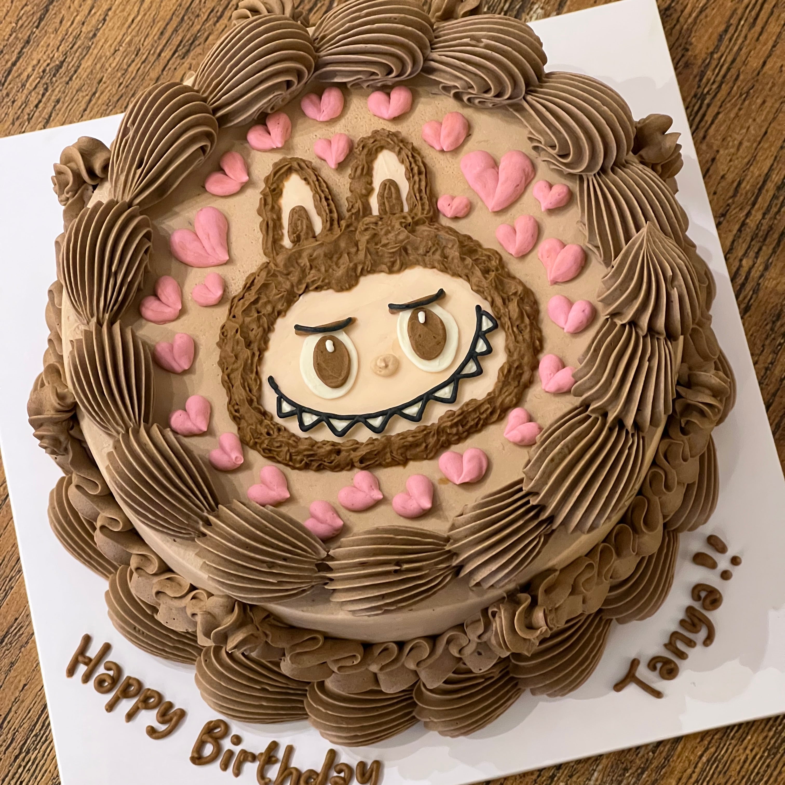 Top view of a round cake featuring Labubu's face, surrounded by brown swirls and pastel pink hearts