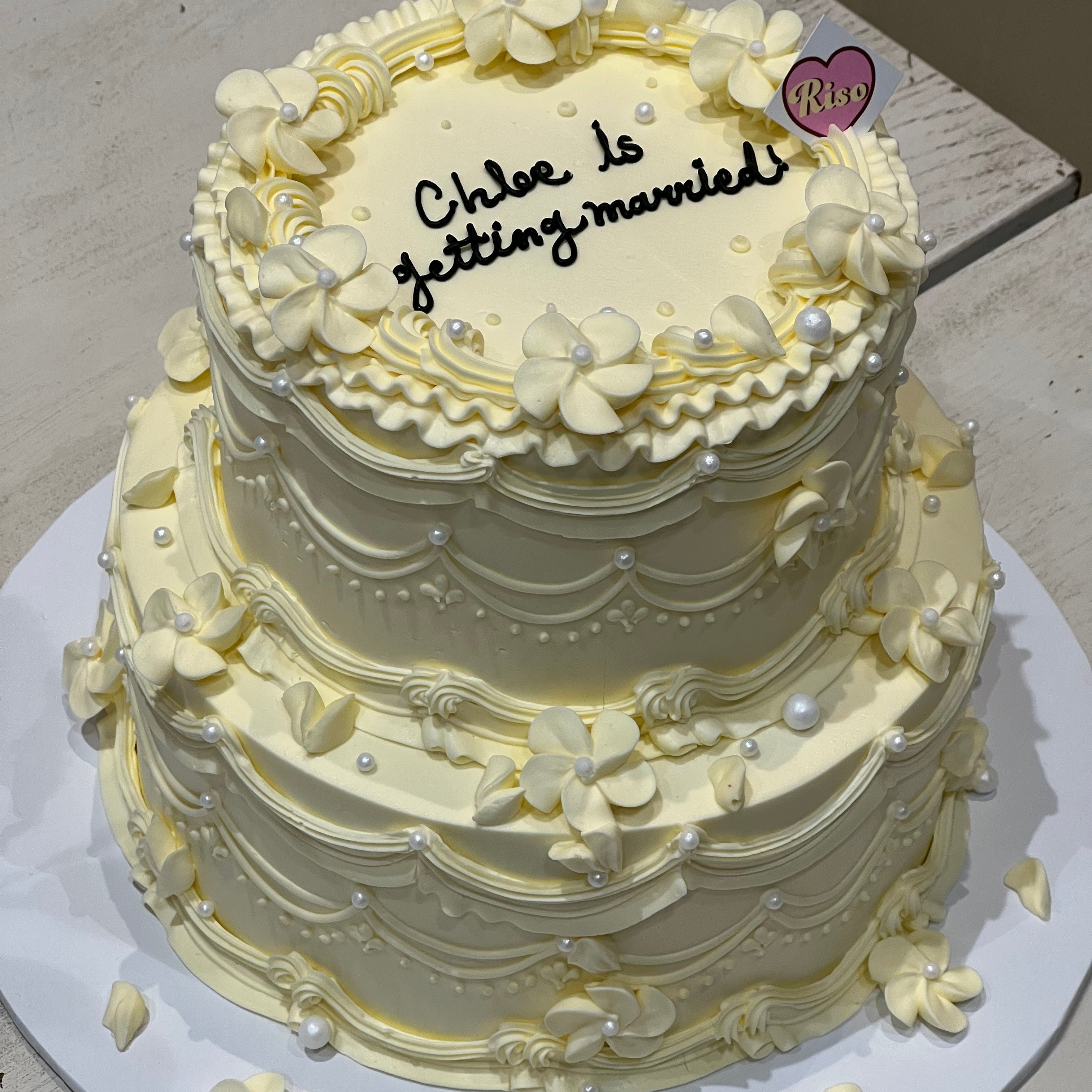 Top view of vintage two-tier engagement cake with cream swirls, floral piping, and heart-shaped Riso topper