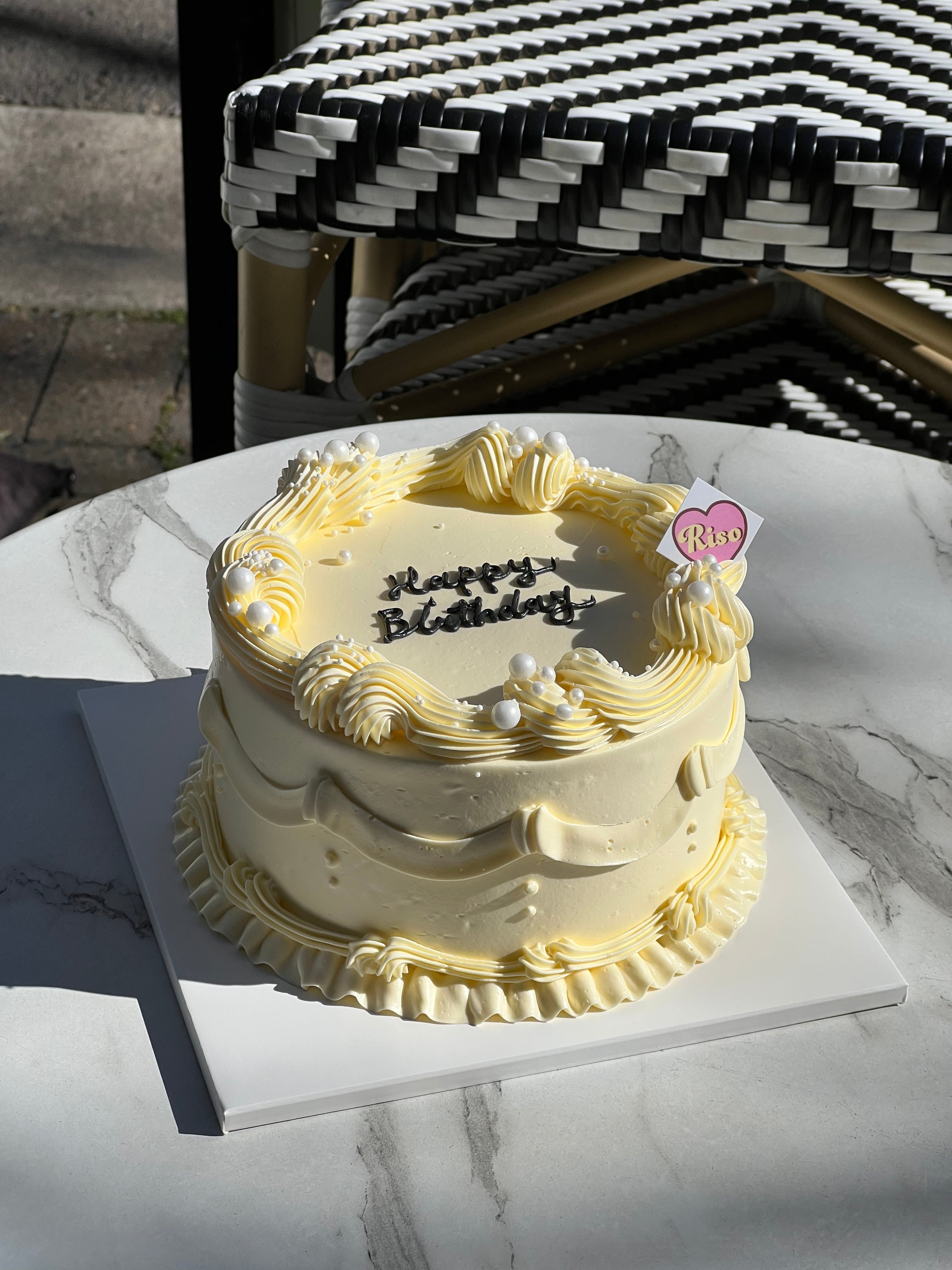 Full view of vintage-style cream birthday cake with piped details, placed on a marble table in natural sunlight