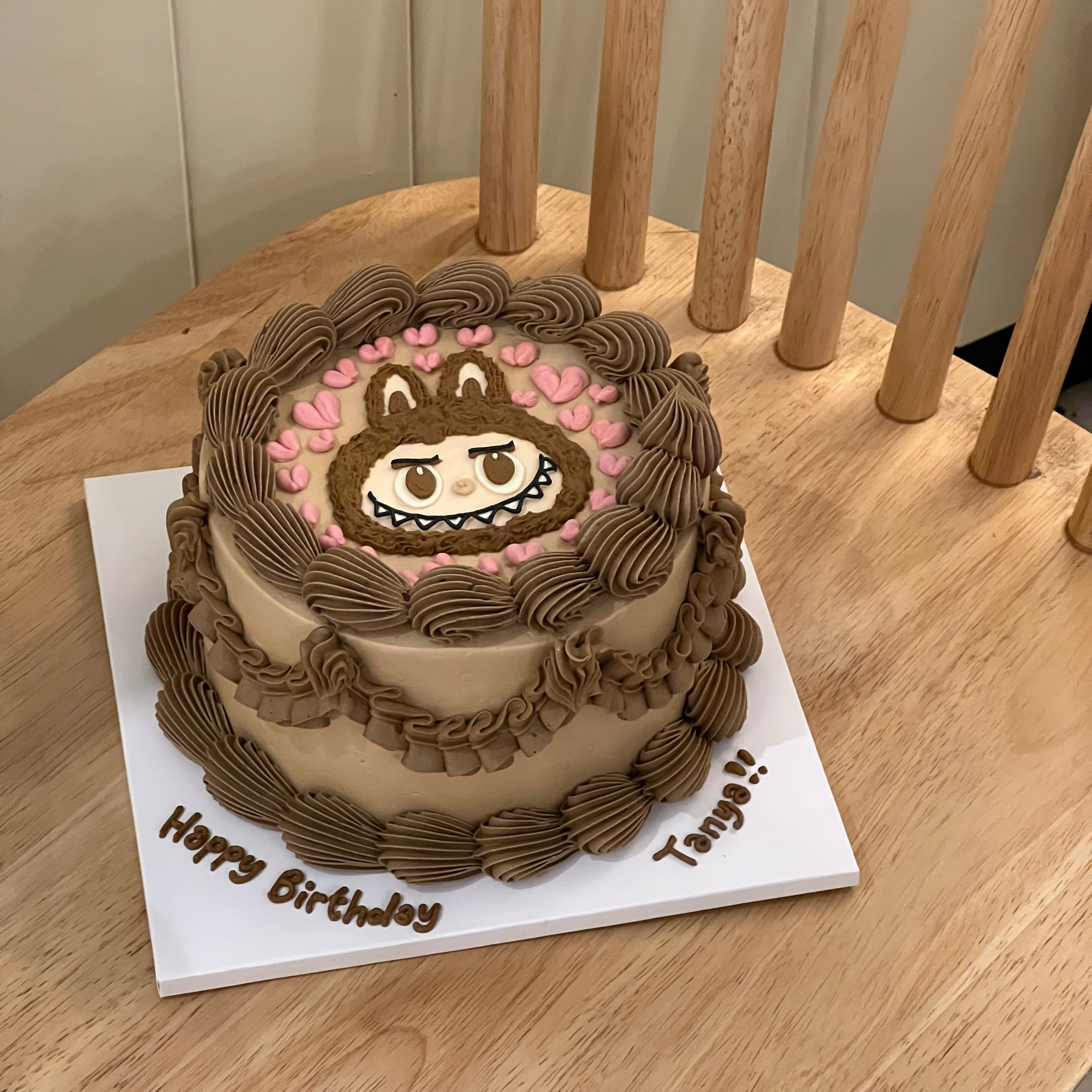Top view of a round cake featuring Labubu's face, surrounded by brown swirls and pastel pink hearts