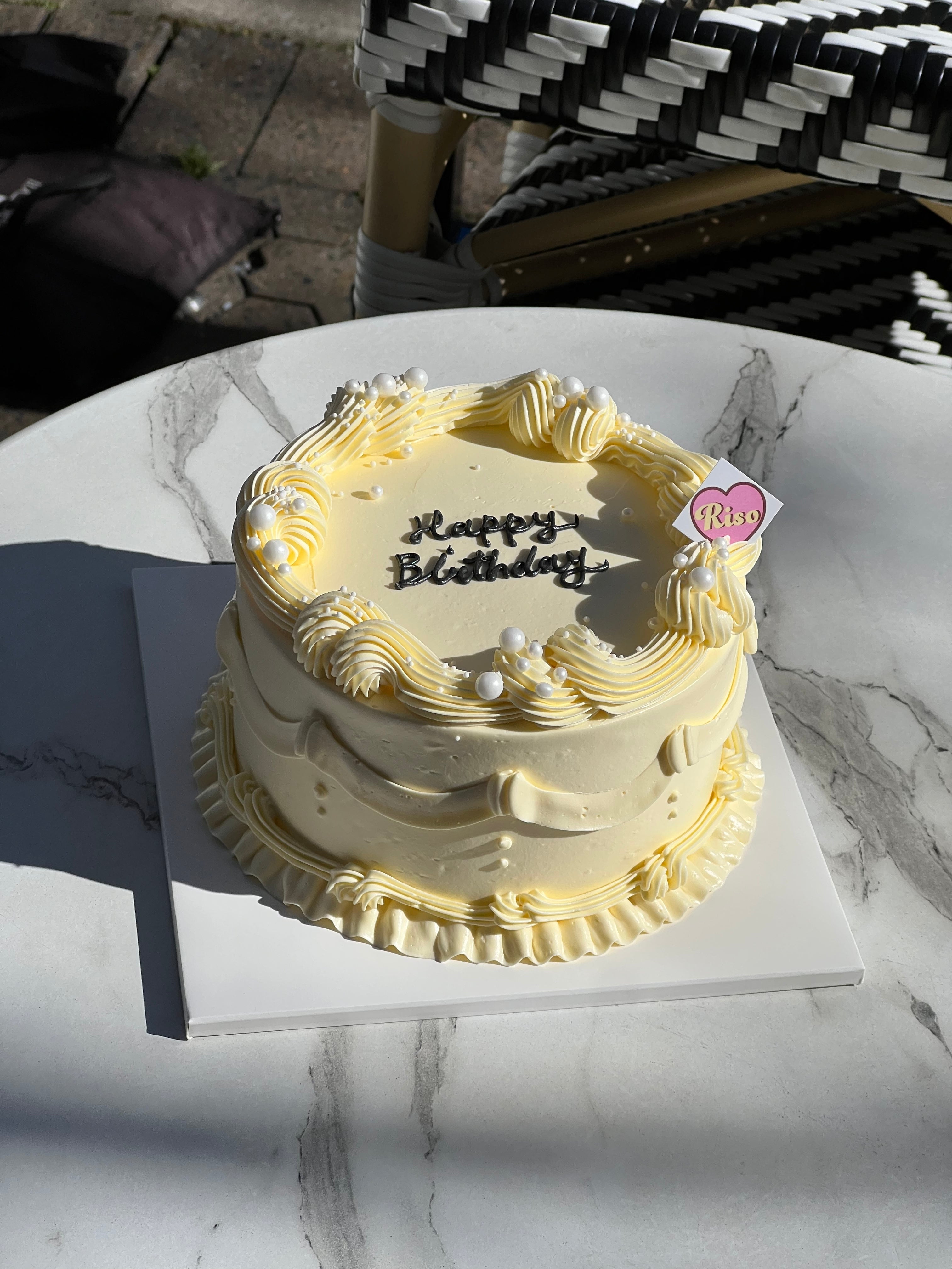 Full view of vintage-style cream birthday cake with piped details, placed on a marble table in natural sunlight
