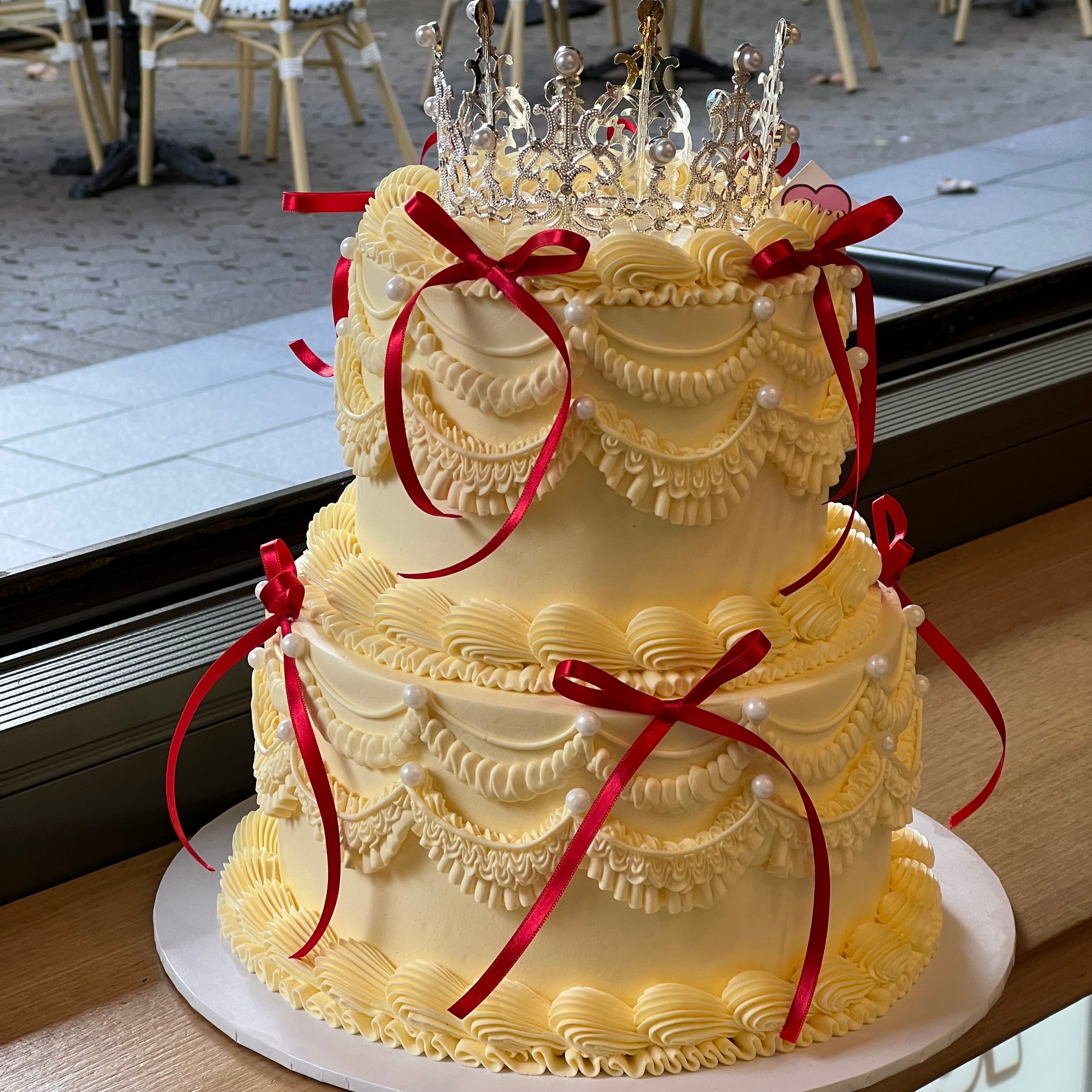 Royal-inspired two-tier cream cake with draped piping, red satin bows, and a glittering crown on top