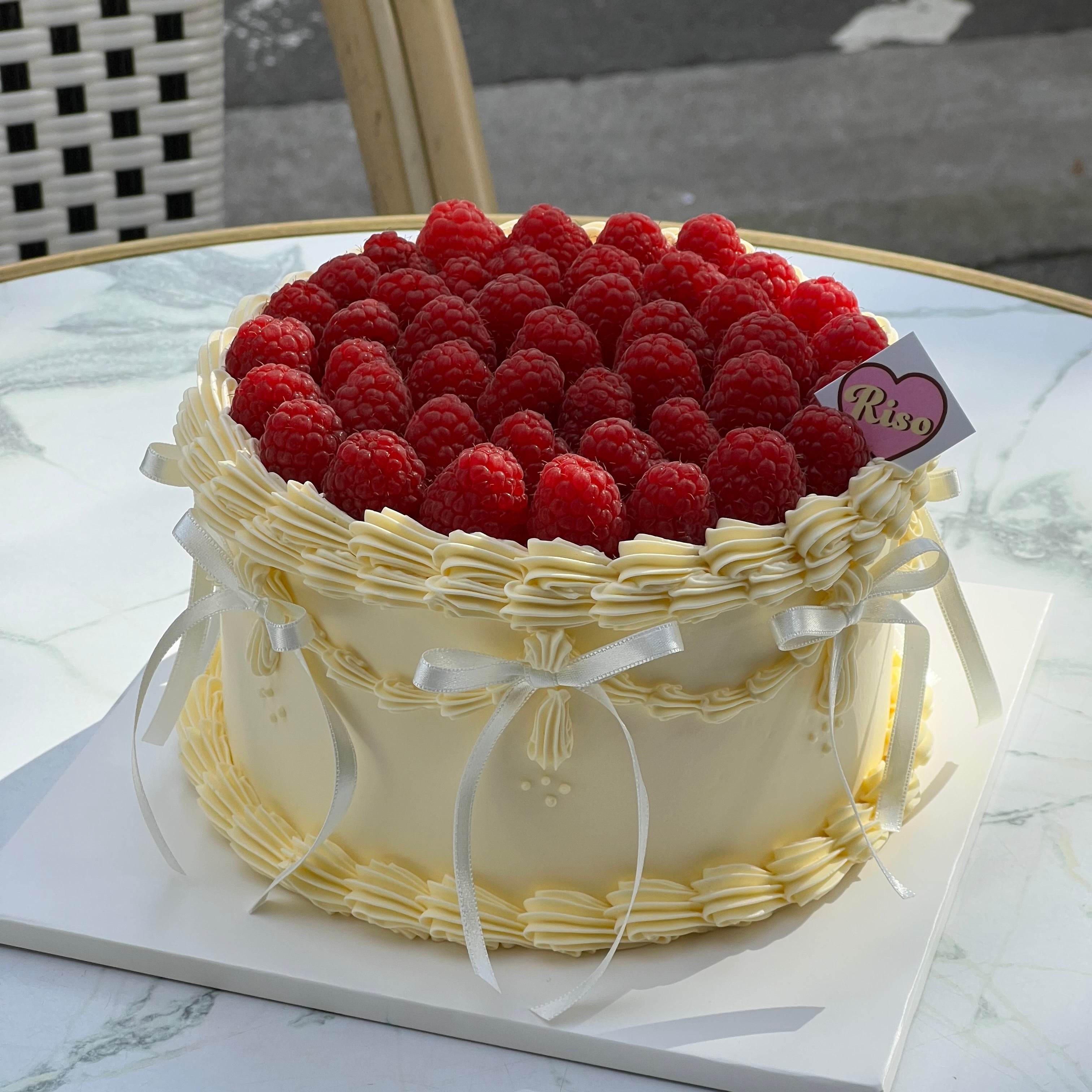A white buttercream cake topped with fresh raspberries and decorated with piped borders and white satin ribbons