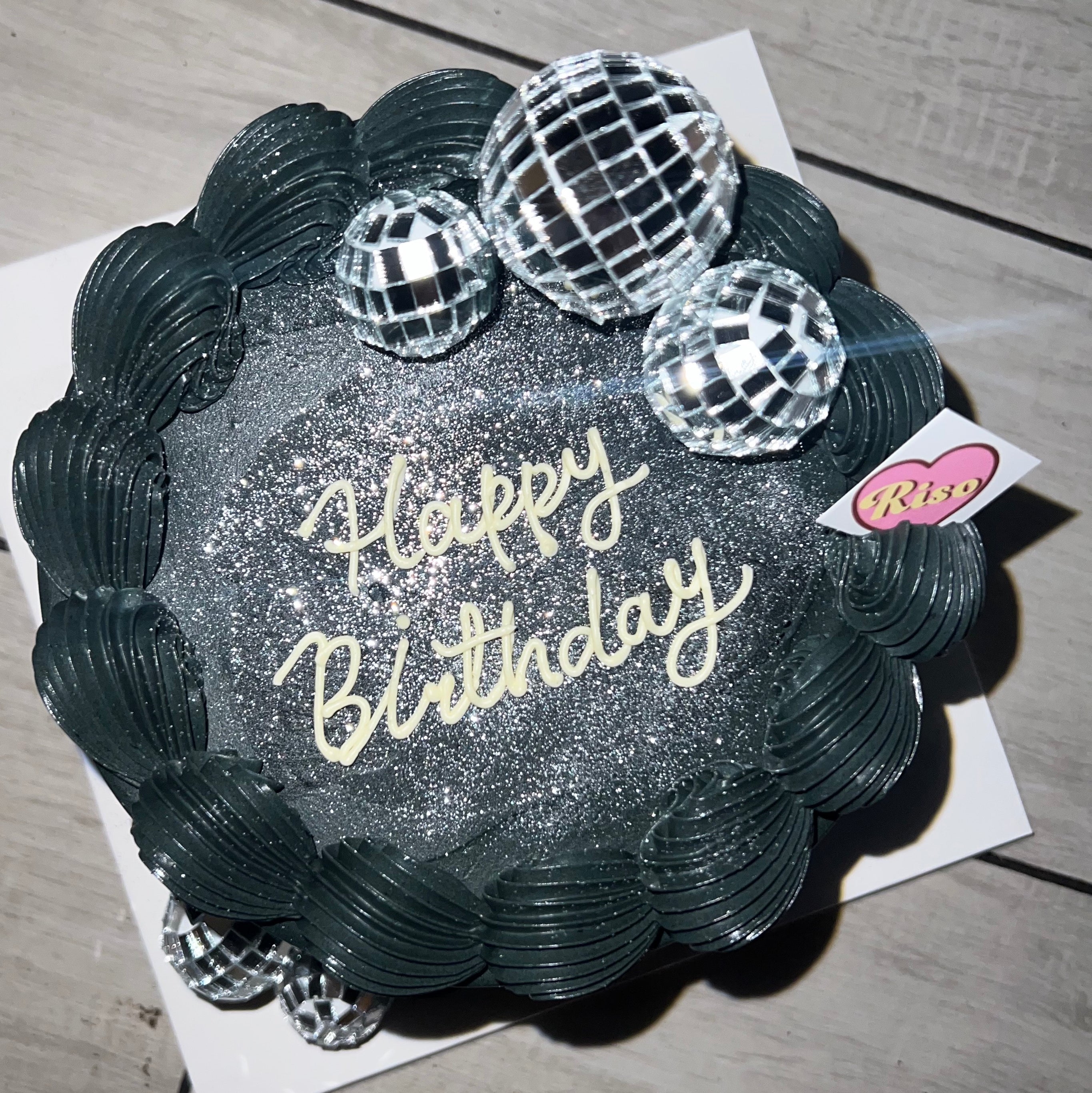 Overhead shot of disco-style black cake with silver shimmer and “Happy Birthday” lettering in cream