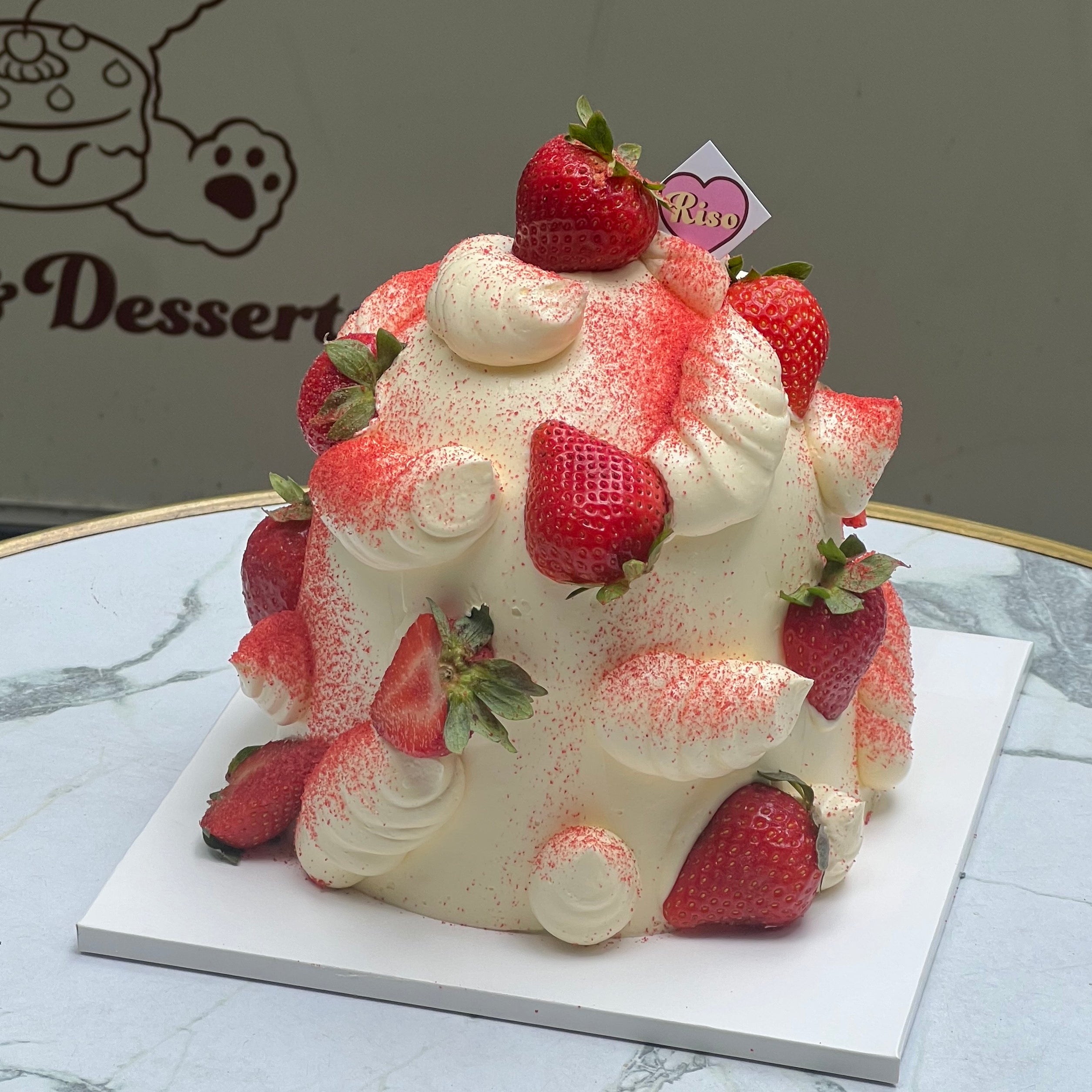 A tall dome-shaped cake covered in whipped cream, decorated with whole strawberries and red powder