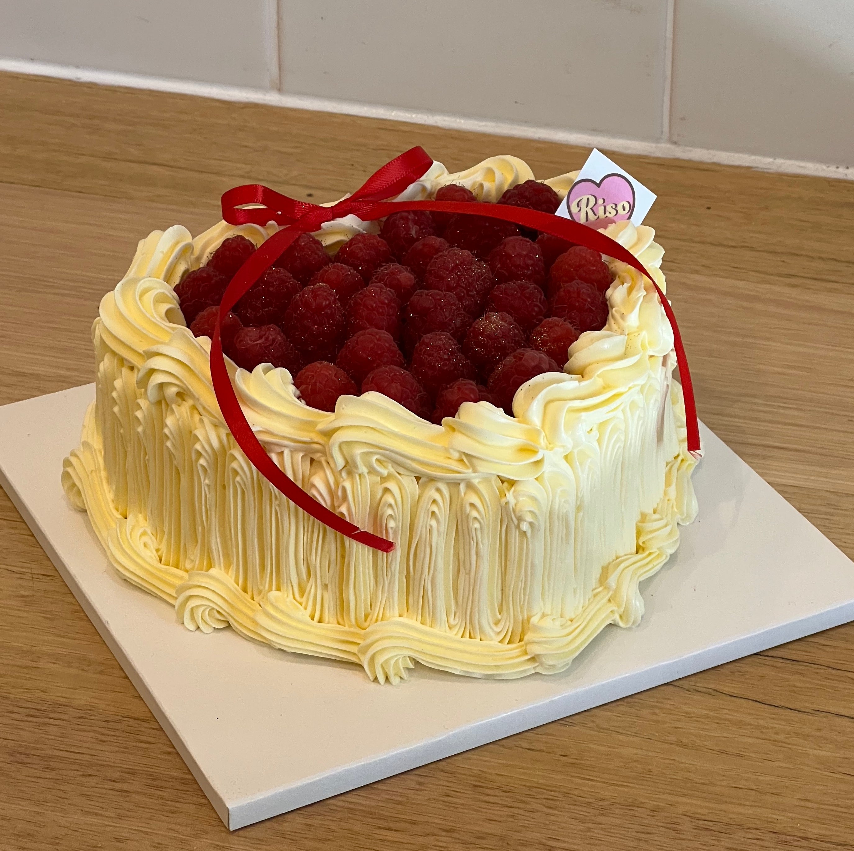 Romantic heart-shaped raspberry cake topped with fresh raspberries and vintage cream piping, finished with a red ribbon — ideal for birthdays, anniversaries, or Valentine’s Day