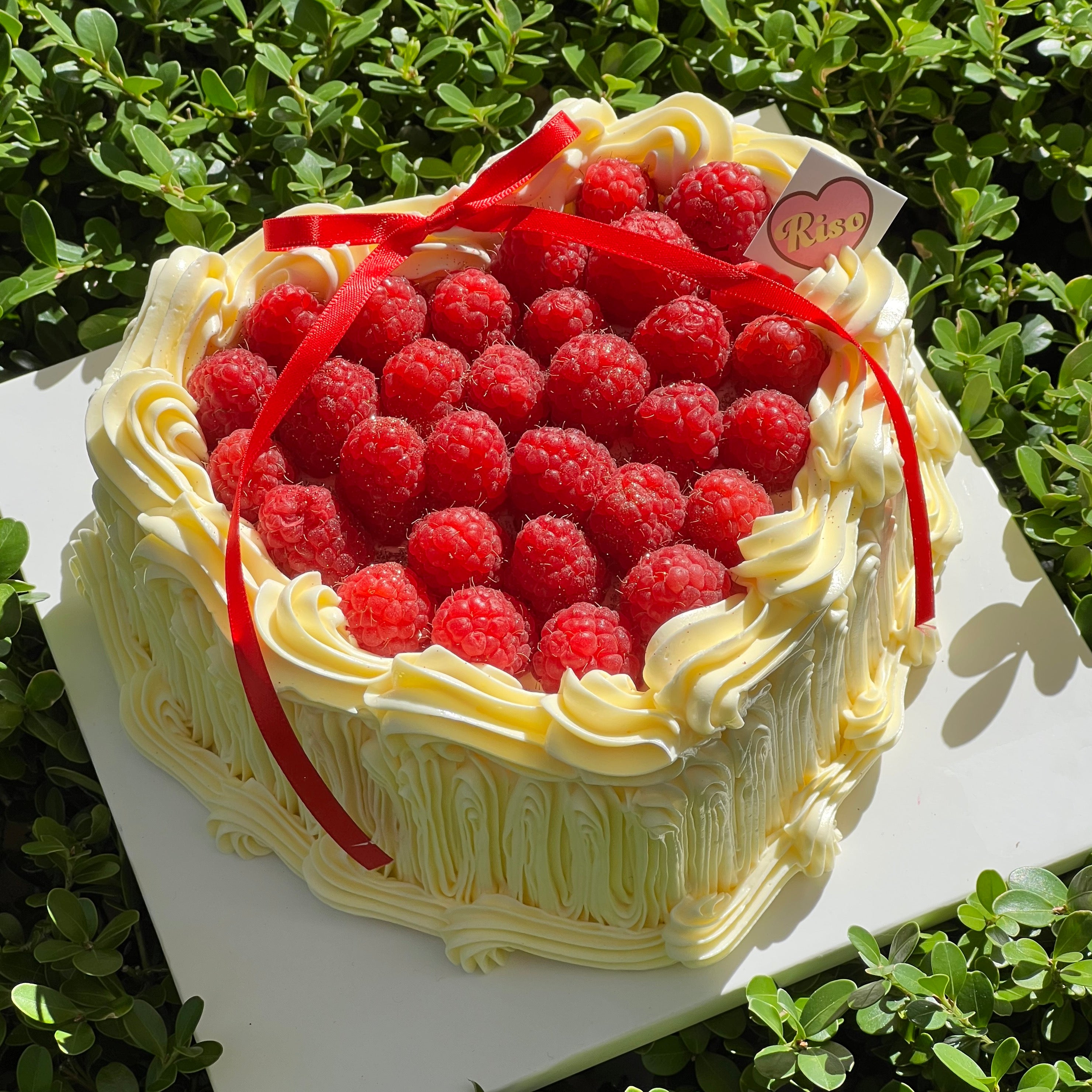 Heart-shaped raspberry cake with piped cream swirls, fresh raspberries, and a red ribbon
