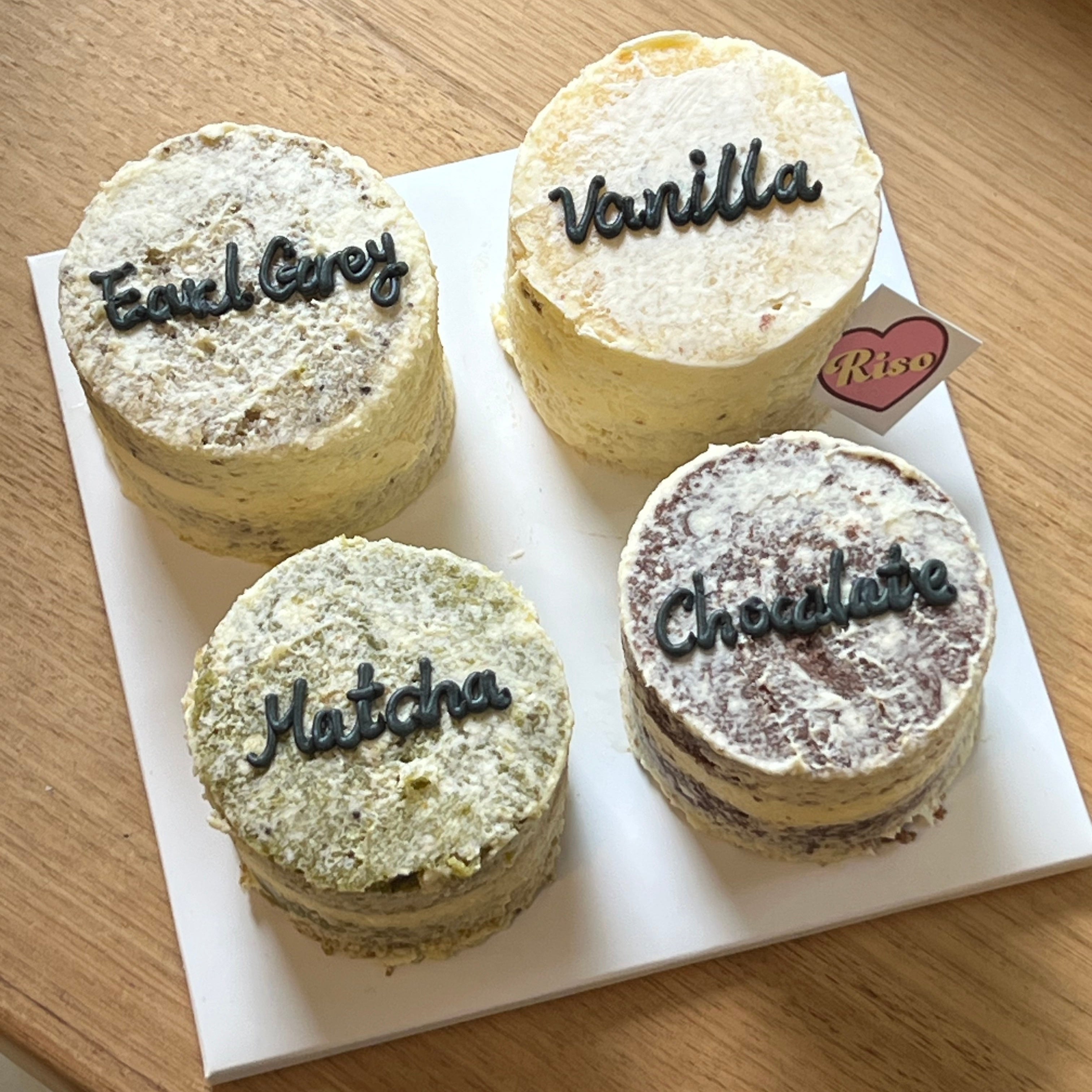 A set of four mini naked cakes labeled Vanilla, Chocolate, Earl Grey, and Matcha, placed beside a pink Riso cake box
