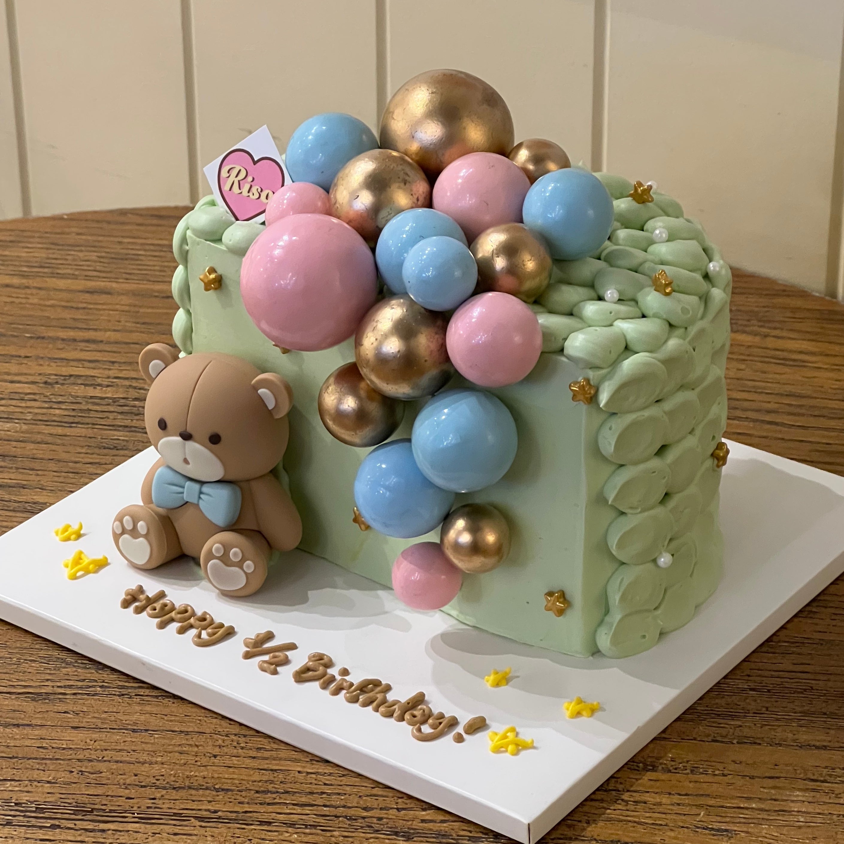 Adorable teddy bear half-birthday cake with pastel pink, blue, and gold balloon toppers, mint green piping, and golden star accents — perfect for 6-month baby celebrations