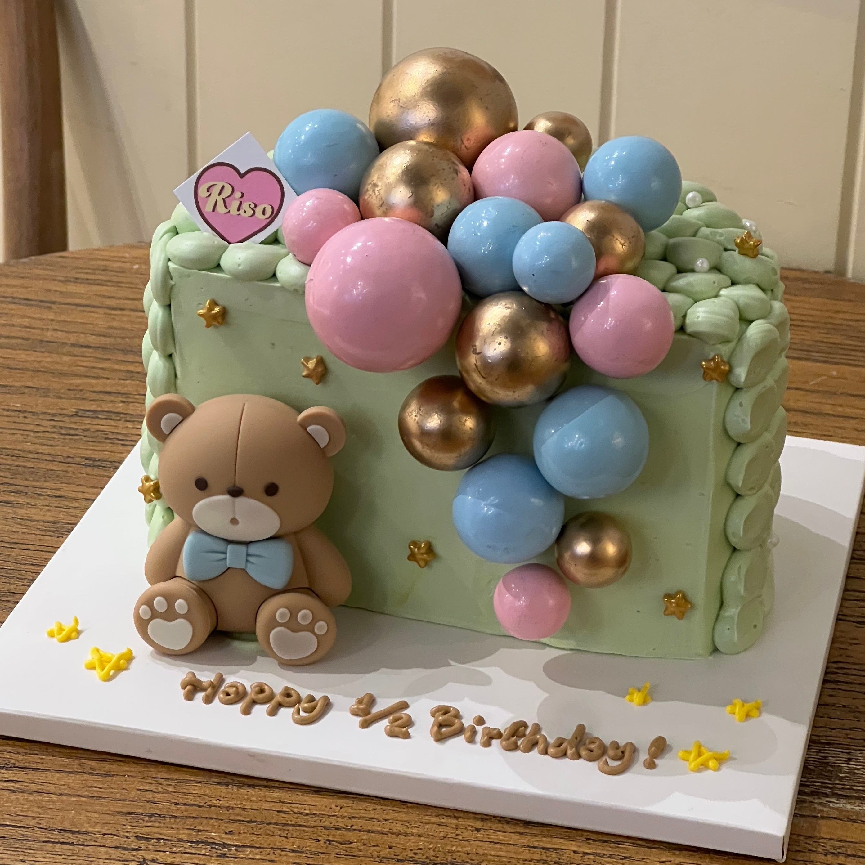 Close-up of a mint-colored ½ birthday cake with piped frosting texture, shiny balloon balls in pink, blue, and gold, and a handmade fondant teddy bear with a blue bow