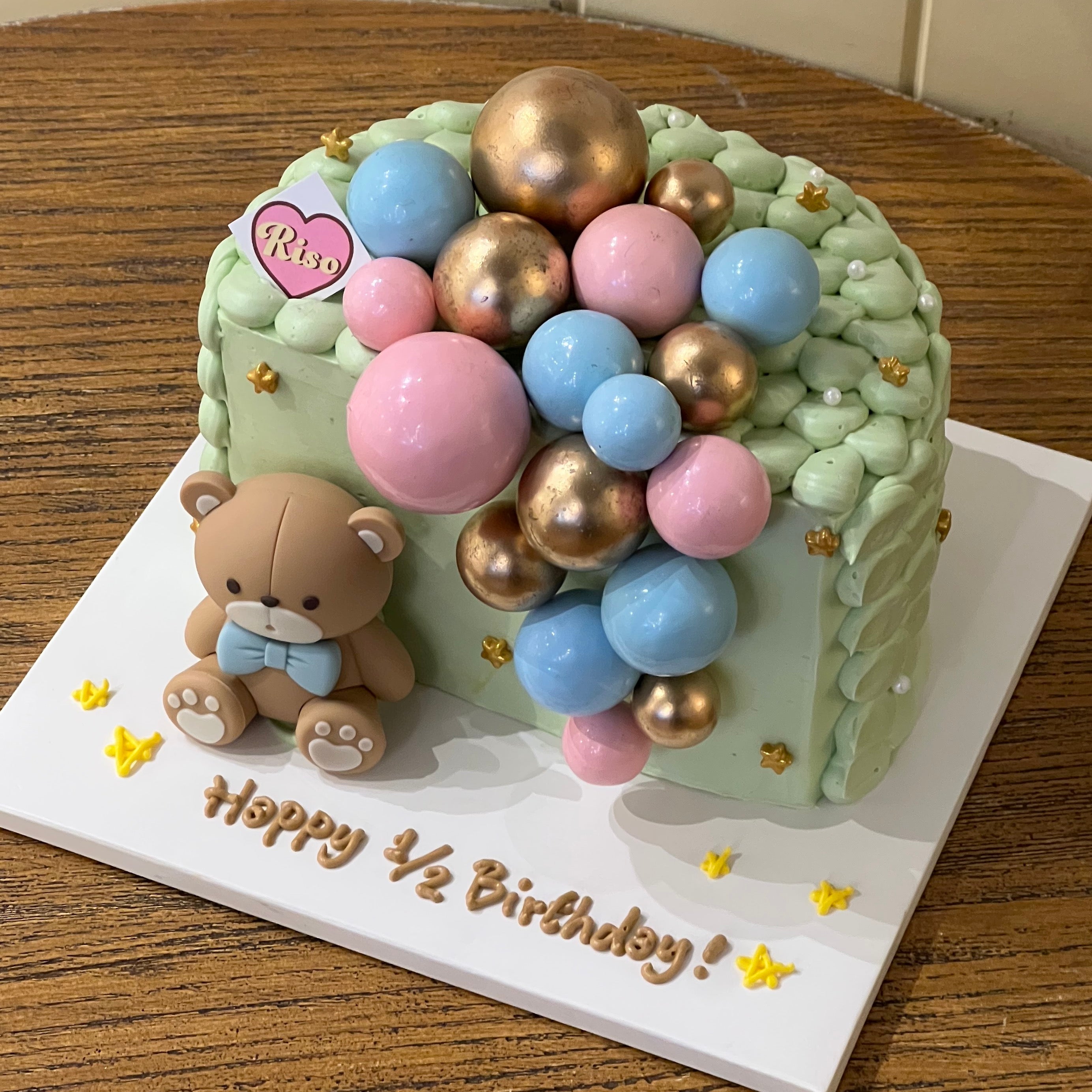 A pastel green half-birthday cake decorated with a fondant teddy bear, colorful balloon toppers, and “Happy ½ Birthday!” written on the base
