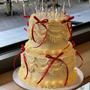 Royal-inspired two-tier cream cake with draped piping, red satin bows, and a glittering crown on top