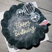 Overhead shot of disco-style black cake with silver shimmer and “Happy Birthday” lettering in cream